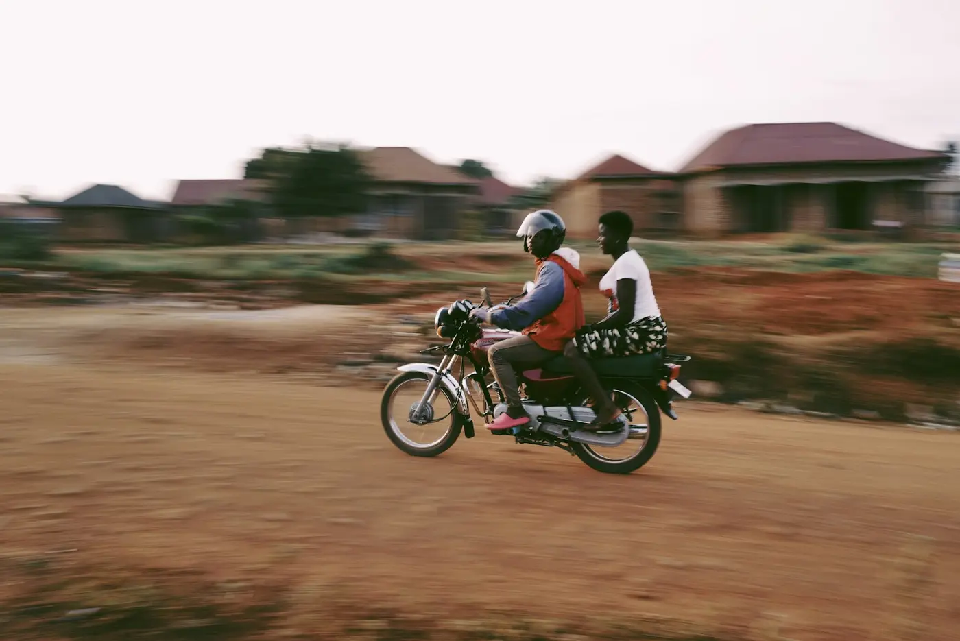 A boda rider carrying a passenger along a red dirt road